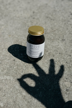 Orchard St Reishi Mushroom powder in an amber glass jar with gold aluminium lid resting on concrete in the sun with the shadow of a hand doing a hand mudra or okay hand pose